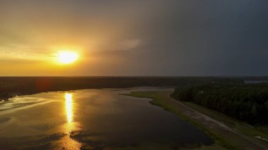 Gün batımında serin bir gölün serin hava manzarası turuncu ve sarı sıcak renkleri yansıtıyor. Yeşillik sularla sınırlıdır. Huzurlu bir atmosfer yaratır..