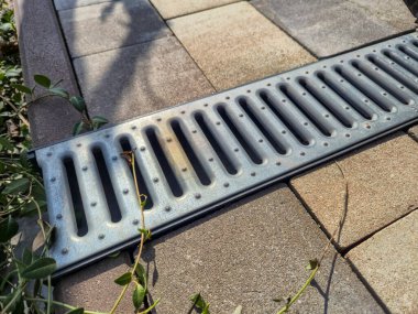 Asfalt kiremitlere döşenmiş metal drenaj ızgarası. Etrafı güneş ışığını alan yeşil bitkilerle çevrili. İnşaat için uygun peyzaj ve ev geliştirme bağlamları.