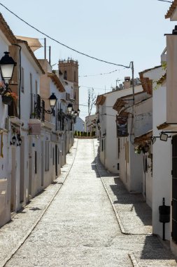Altea İspanya - 17 Temmuz 2025: Altea 'da dar bir güneş ışığı sokağı geleneksel beyazlatılmış binalar fenerler ve uzak saat kulesine uzanan hafif bir yamaç içerir..