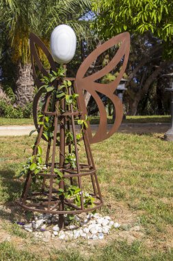 Alicante Spain - 13 Temmuz 2025: Artistik metal heykel, arka planda palmiye ağaçları olan canlı bir bahçe manzarasına yerleştirilmiş yaprak motifli bir meleğe benziyor.