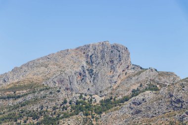 İspanya 'nın Guadalest bölgesindeki Puig Campana dağının çarpıcı manzarası açık mavi gökyüzünün altında engebeli tepeler ve yeşil bitkiler sergiliyor. Seyahat ve doğa meraklıları için ideal..