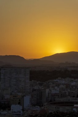 Güneş ufukta batarken Alicante 'nin muhteşem günbatımı manzarası ikonik ufuk çizgisini ve etrafındaki dağları gösteriyor. Gökyüzünde sıcak turuncu renkler döküyor..