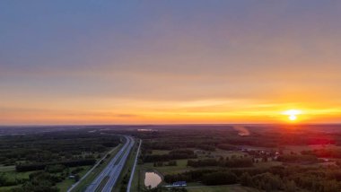 Gün batımında sakin manzarayı görüntüleyen hava manzarası. Bereketli yeşillik ve alacakaranlığın ılık tonlarını yansıtan sakin bir göletin yanında dolambaçlı bir otoyol..