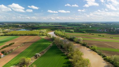 Bereketli yeşil tarlaların ve parlak mavi gökyüzünün altında kıvrımlı bir nehrin havadan görünüşü kabarık bulutların bahar zamanı kırsal manzaraların güzelliğini gözler önüne seriyor..