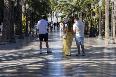 Alicante Spain - July 17 2025: A vibrant scene along a palm-lined promenade in Alicante featuring three people engaged with their smartphones enjoying a sunny day.