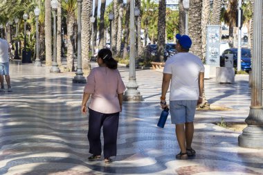 Alicante Spain - July 17 2025: Two people walking along a palm-lined promenade on a sunny day enjoying leisure time together in a vibrant coastal city.