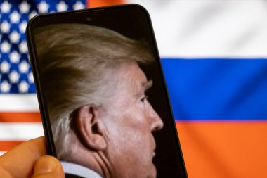 Warsaw Poland - September 17 2025: A close-up of an individual holding a smartphone displaying a profile of Donald Trump with the American and Russian flags in the background symbolizing global politics.