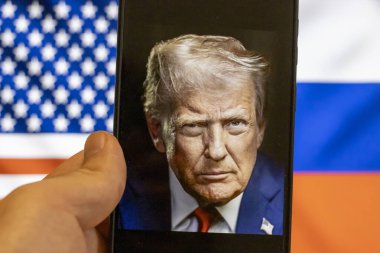 Warsaw Poland - September 17 2025: A close-up of a hand holding a smartphone displaying a portrait of Donald Trump with American and Russian flags in the background highlighting a political theme.