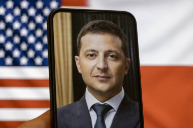 Warsaw Poland - September 17 2025: A person holding a smartphone displaying a portrait of Volodymyr Zelensky with American and Polish flags in the background symbolizes unity amid the Ukraine war concept.