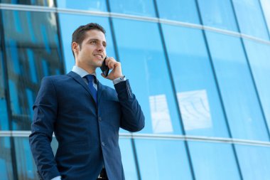 Mutlu genç işadamı dışarıda cam binada telefonla konuşuyor.
