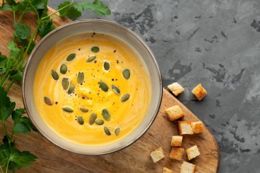 Tahta tahtada servis edilen balkabağı çorbası, kraker ve gri arka planda taze yeşil. Üst görünüm