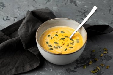 Derin kasede kabak çorbası, tohumlar ve kaşık ile servis edilen gri arka planda peçete.. 