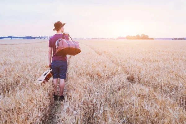 若い息子の家出 ロマンチックな旅行バック グラウンド 日没のフィールドで歩いているギターと道路のバッグを持つ男 — ストック写真