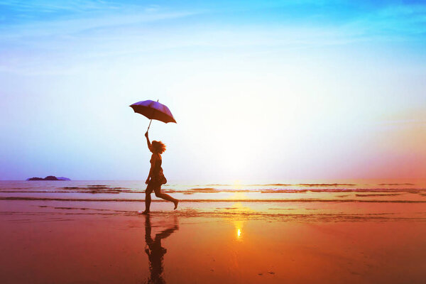 silhouette of happy carefree girl with umbrella jumping on the beach at sunset, freedom and joy concept