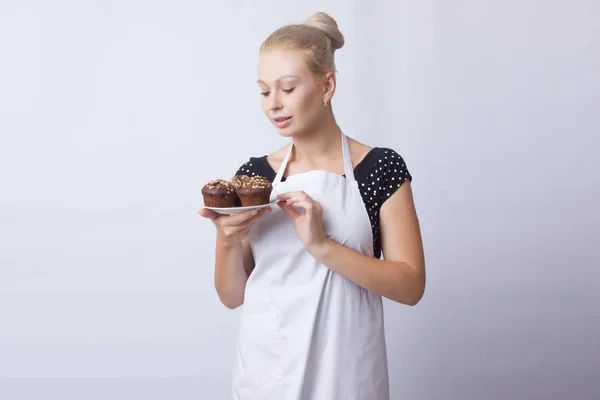 A happy blond woman in a white apron shows a chocolate cupcakes on a white plate. Cook. A housewife