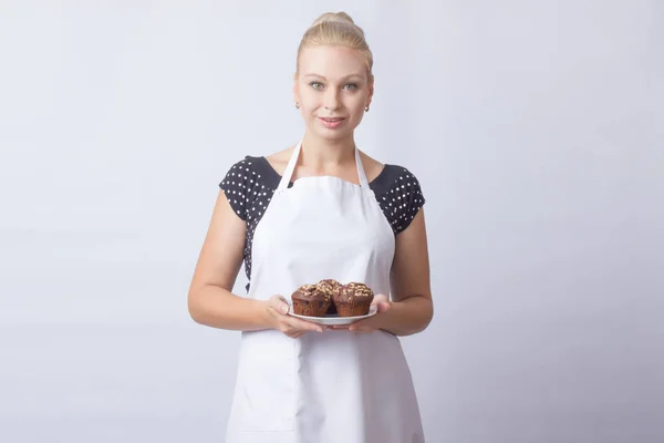 A happy blond woman in a white apron shows a chocolate cupcakes on a white plate. Cook. A housewife