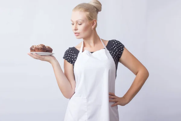 Mutlu sarışın bir kadın beyaz bir önlük beyaz bir plaka üzerinde bir çikolata cupcakes gösterir. Aşçı. Bir ev kadını