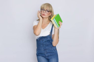 Güzel bir sarışın genç kadın mavi kot tulum ve kitaplar onun elinde tutan gri arka plan üzerinde gözlük Studio portresi