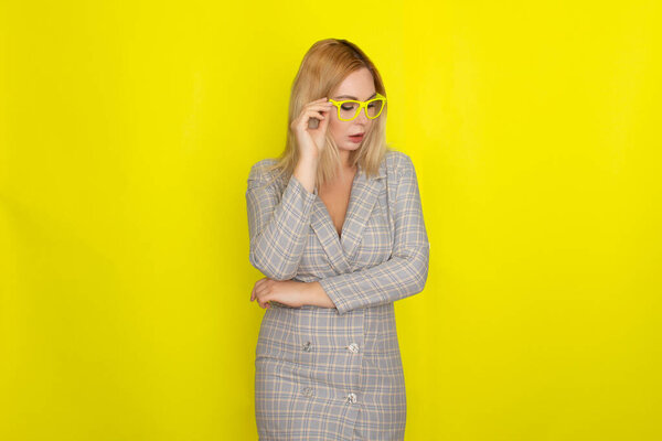 Attractive blonde woman in plaid jacket dress over yellow background wearing yellow glasses