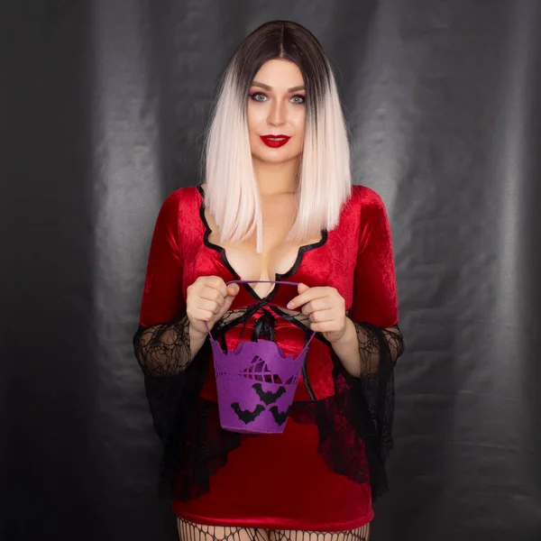 若いです美しいですブロンド女性で赤吸血鬼ドレス保持していますハロウィンパープルバッグとともにバットのためのお菓子 — ストック写真