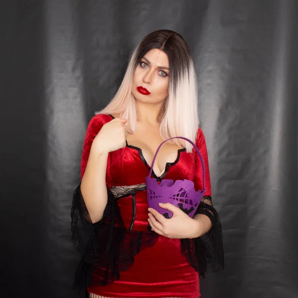 若いです美しいですブロンド女性で赤吸血鬼ドレス保持していますハロウィンパープルバッグとともにバットのためのお菓子 — ストック写真