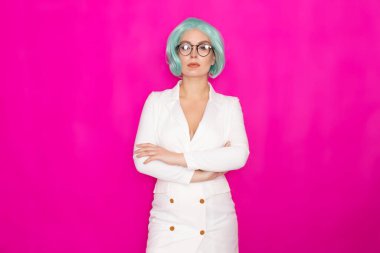 Kısa mavi saçlı, gözlüklü, beyaz iş elbisesi giyen, stüdyoda parlak pembe bir arka planda poz veren güzel bir genç kadın.