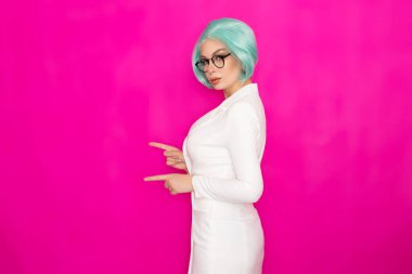 Kısa mavi saçlı, gözlüklü, beyaz iş elbisesi giyen, stüdyoda parlak pembe bir arka planda poz veren güzel bir genç kadın.