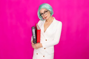 Kısa mavi saçlı, beyaz iş elbisesi içinde gözlük takan güzel bir genç kadın elinde iki kalın klasör tutuyor.