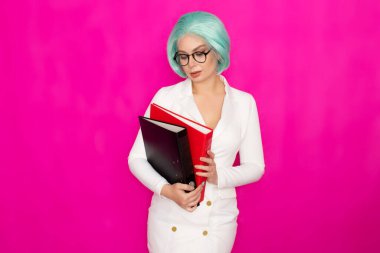 Kısa mavi saçlı, beyaz iş elbisesi içinde gözlük takan güzel bir genç kadın elinde iki kalın klasör tutuyor.