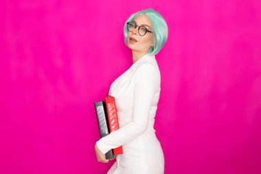 Kısa mavi saçlı, beyaz iş elbisesi içinde gözlük takan güzel bir genç kadın elinde iki kalın klasör tutuyor.