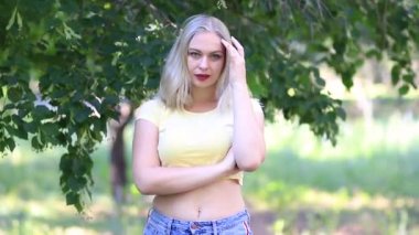 Yaz şortlu sarışın kadın ve yeşil çimen ve ağaçların arka planında açık havada poz veren sarı kısa tişörtlü.
