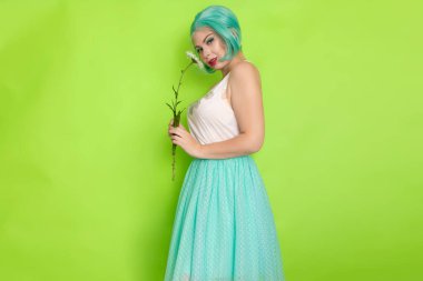 Stüdyo portresi, beyaz bluzlu, açık mavi etekli, yeşil arka planda büyük bir papatya çayı tutan güzel, nane rengi saçlı bir kadın.