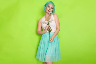 Stüdyo portresi, beyaz bluzlu, açık mavi etekli, yeşil arka planda büyük bir papatya çayı tutan güzel, nane rengi saçlı bir kadın.