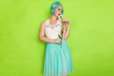 Stüdyo portresi, beyaz bluzlu, açık mavi etekli, yeşil arka planda büyük bir papatya çayı tutan güzel, nane rengi saçlı bir kadın.