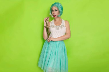 Stüdyo portresi, beyaz bluzlu, açık mavi etekli, yeşil arka planda büyük bir papatya çayı tutan güzel, nane rengi saçlı bir kadın.