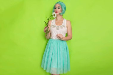Stüdyo portresi, beyaz bluzlu, açık mavi etekli, yeşil arka planda büyük bir papatya çayı tutan güzel, nane rengi saçlı bir kadın.