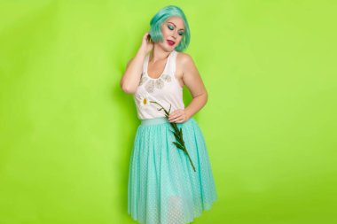 Stüdyo portresi, beyaz bluzlu, açık mavi etekli, yeşil arka planda büyük bir papatya çayı tutan güzel, nane rengi saçlı bir kadın.