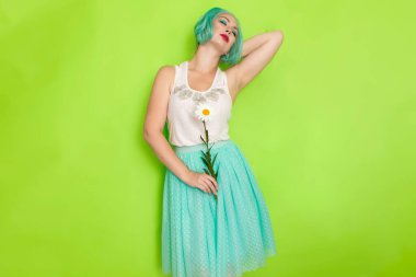 Stüdyo portresi, beyaz bluzlu, açık mavi etekli, yeşil arka planda büyük bir papatya çayı tutan güzel, nane rengi saçlı bir kadın.