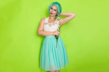 Stüdyo portresi, beyaz bluzlu, açık mavi etekli, yeşil arka planda büyük bir papatya çayı tutan güzel, nane rengi saçlı bir kadın.