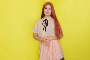 Pembe bluzlu ve etekli, sarı arka planda poz veren genç, güzel, kızıl saçlı bir kadın. Yaz konsepti