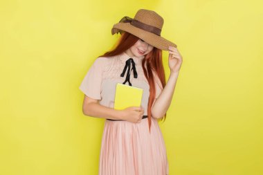 Sarı arka planda saman şapkalı kızıl saçlı kadın elinde kitap tutuyor, stüdyo, tarz, yaz konsepti.