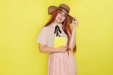 Sarı arka planda saman şapkalı kızıl saçlı kadın elinde kitap tutuyor, stüdyo, tarz, yaz konsepti.