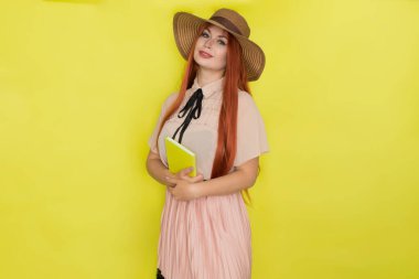 Sarı arka planda saman şapkalı kızıl saçlı kadın elinde kitap tutuyor, stüdyo, tarz, yaz konsepti.