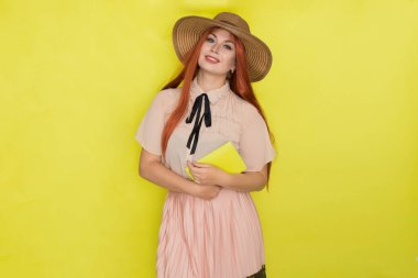 Sarı arka planda saman şapkalı kızıl saçlı kadın elinde kitap tutuyor, stüdyo, tarz, yaz konsepti.