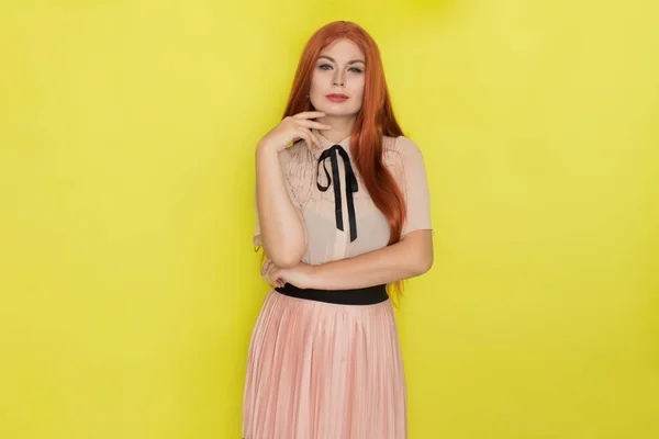 Single gorgeous young red haired woman in pink blouse and skirt posing over yellow background. Summer concept