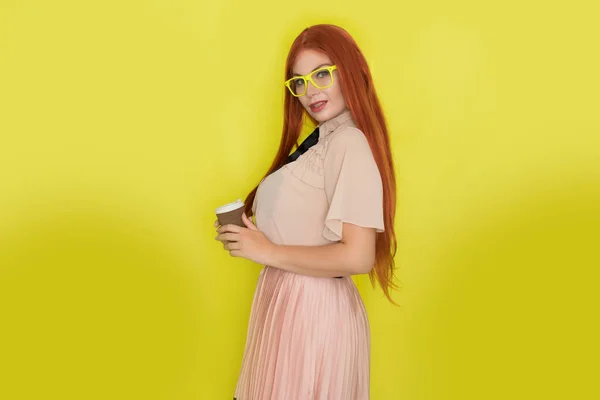 黄色の背景 スタジオ スタイル上のコーヒーや紅茶のカップを保持眼鏡やオフィス服を持つ赤い髪の女性 — ストック写真