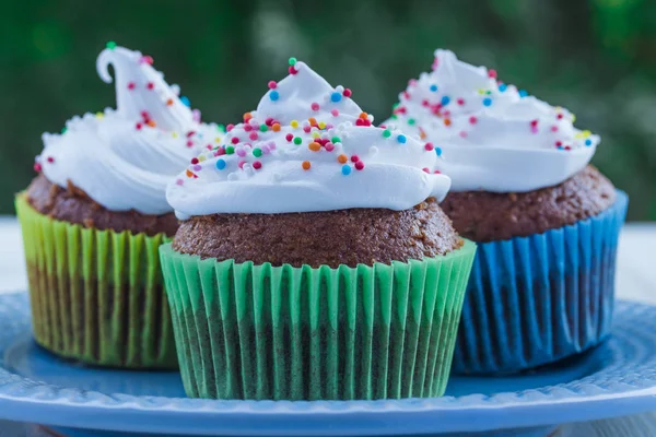 Yeşil yapraklarıbir arka plan üzerinde ahşap bir masa üzerinde beyaz krem ve renkli serpilir ile Lezzetli muffins.