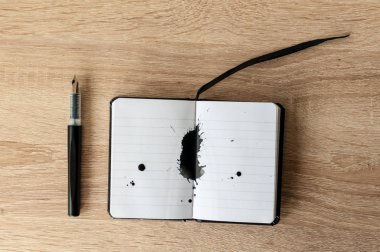 Dolma kalem üstünde bir defter. Siyah mürekkep sıçrama. Ahşap masa. Kopya alanı.
