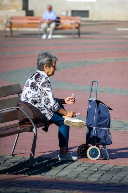 Timisoara, Romanya - 05 Eylül 2020: Yaşlı bir kadın bankta oturuyor ve sigara içiyor. Gerçek insanlar..