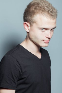 Kısa sarı saçları ve siyah t-shirt giyen ve gri arka plan üzerinde poz yüzündeki çiller ile komik gülümseyen genç adam portresi. Hippi tarzı. Stüdyo vurdu